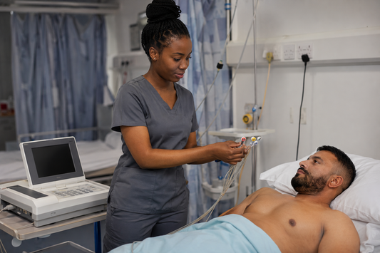 Student nurse in grey scrubs preparing ECG leads beside an adult patient in a hospital bed before recording a 12-lead ECG.